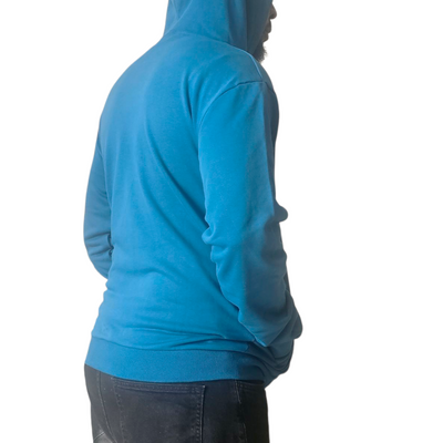 Person wearing a blue hoodie with a small yellow geometric logo on the left chest, hands in front pocket, and hood up, standing against a plain white background
