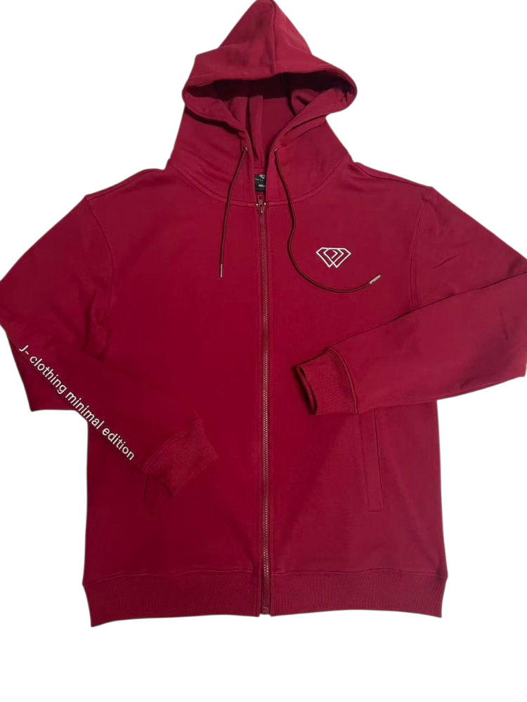 A man with short dark hair and a beard walks on a city street wearing a red J-clothing premium cotton hoodie, blue jeans, and white sneakers. The background is slightly blurred, showing buildings and outdoor seating.