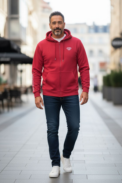 A man with short dark hair and a beard walks on a city street wearing a red J-clothing premium cotton hoodie, blue jeans, and white sneakers. The background is slightly blurred, showing buildings and outdoor seating.