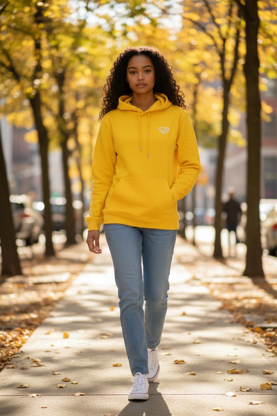 A woman with curly hair walks confidently down a tree-lined sidewalk on a sunny autumn day, wearing a J-clothing premium cotton yellow pullover hoodie and classic blue jeans that elevate her streetwear style.