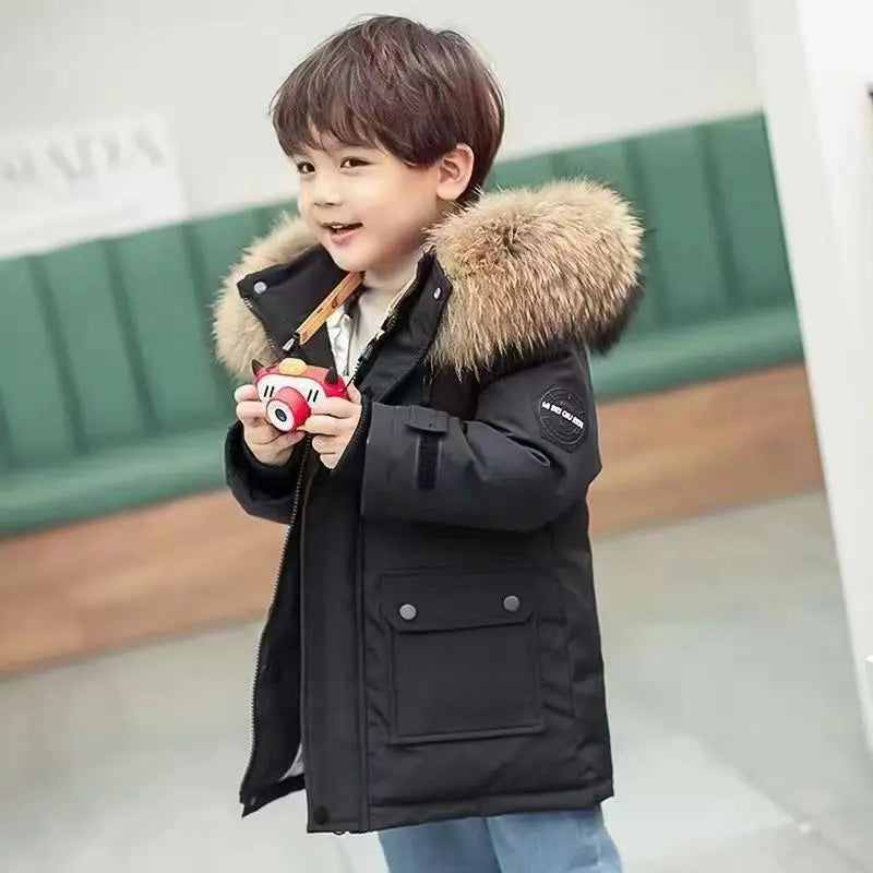 A smiling young child is wearing a Jstoremart boys' winter coat with a fur-trimmed hood while standing indoors. The hooded down jacket is buttoned up, and the setting appears to be a warm, well-lit room.