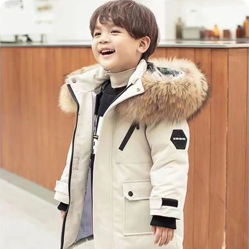 A smiling young child is wearing a Jstoremart boys' winter coat with a fur-trimmed hood while standing indoors. The hooded down jacket is buttoned up, and the setting appears to be a warm, well-lit room.