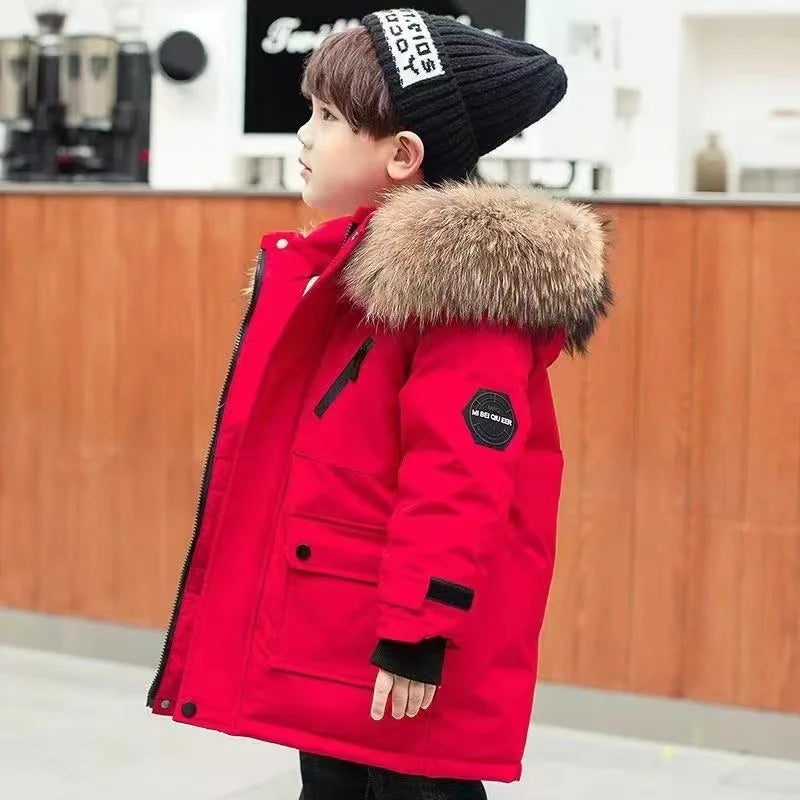 A young child sporting a Jstoremart red coat with a fur-lined hood and black beanie stands indoors. This stylish boys' winter jacket by Jstoremart features multiple pockets and black accents. The child looks sideways, framed by wooden panels and some equipment in the background.